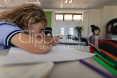 Side view of schoolgirl leaning on desk and writing on notebook in classroom