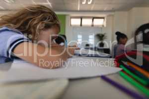 Side view of schoolgirl leaning on desk and writing on notebook in classroom