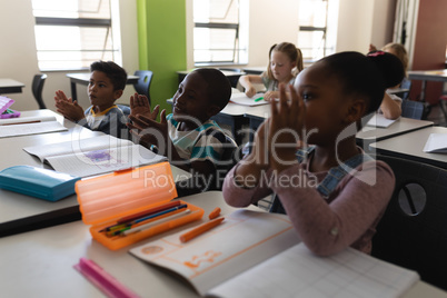 Side view of schoolkids applauding and sitting at desk in classroom