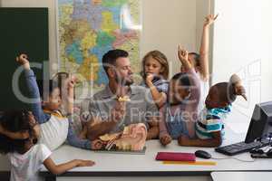 Front view of schoolkids raising hands while teacher teaching anatomy at desk in classroom