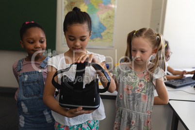 Front view of schoolgirl with classmates holding and looking virtual reality headset in classroom