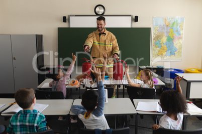 Schoolkids raising hands while male firefighter teaching about fire safety in classroom