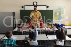 Schoolkids raising hands while male firefighter teaching about fire safety in classroom
