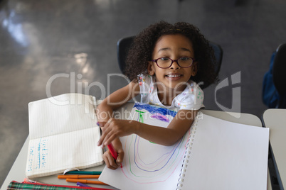 High angle view of schoolgirl drawing in book while looking at camera in classroom
