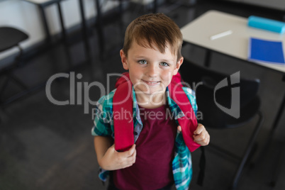 High angle view of happy schoolboy with schoolbag looking at camera in classroom