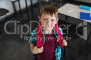 High angle view of happy schoolboy with schoolbag looking at camera in classroom