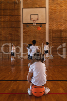 Rear view of schoolgirl sitting on basketball at basketball court