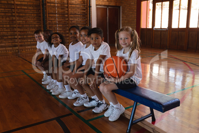 Side view of schoolkids with basketball sitting on bench looking at camera