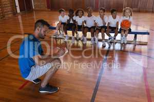 Basketball coach writing on clipboard and schoolkids sitting on bench at basketball court