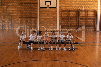 Front view of schoolkids with basketball sitting on bench at basketball court