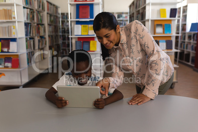 Female teacher teaching schoolboy on digital tablet at table in school library