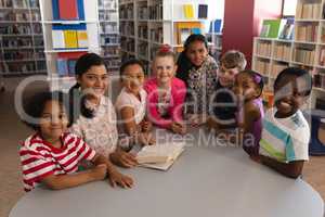 Happy female teacher and schoolkids looking at camera while studying at table in school library