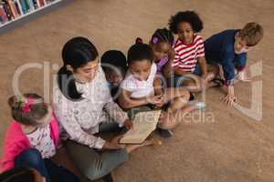 Female teacher teaching schoolkids and sitting on floor of school library