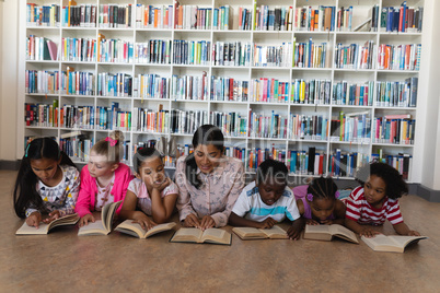 Female teacher and schoolkids reading a book while lying on floor of school library