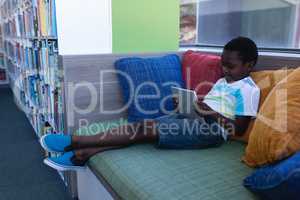 Schoolboy playing on digital tablet while sitting on couch in school library