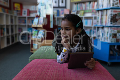 Thoughtful schoolgirl with hand on chin holding digital tablet and looking away in school library