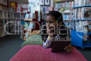 Thoughtful schoolgirl with hand on chin holding digital tablet and looking away in school library