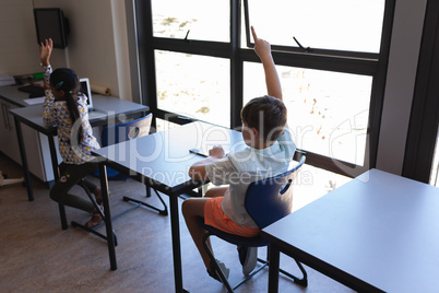 Schoolkid raising their hand while sitting at desk in classroom