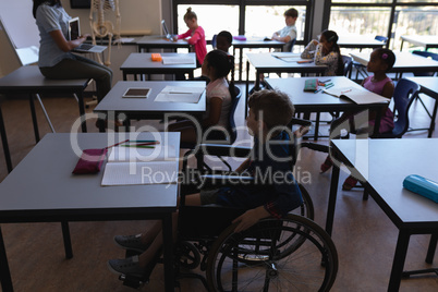 Disable schoolboy with classmates studying and sitting at desk in classroom