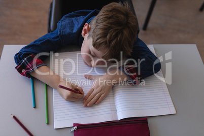 Schoolboy writing on notebook and sitting at desk in classroom