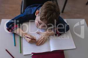 Schoolboy writing on notebook and sitting at desk in classroom
