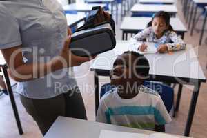 Female teacher wearing virtual reality headset to schoolboy at desk in classroom