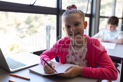 Schoolgirl writing on notebook and looking at camera at desk in classroom
