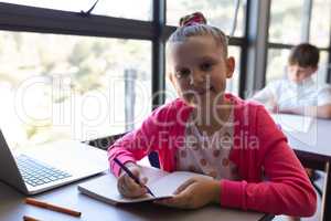 Schoolgirl writing on notebook and looking at camera at desk in classroom