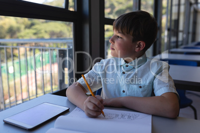 Schoolboy looking away while drawing on notebook at desk in classroom