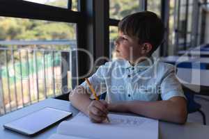Schoolboy looking away while drawing on notebook at desk in classroom