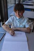 Schoolboy drawing on notebook and looking at camera at desk in classroom