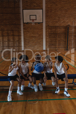 Schoolkids talking with each other and sitting on bench at basketball court