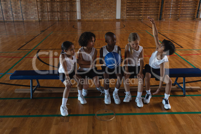 Schoolkids talking with each other and sitting on bench at basketball court