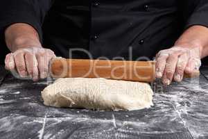 chef in a black tunic rolls a dough for a round pizza