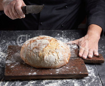 male chef hand holds knife over a whole round baked loaf of brea