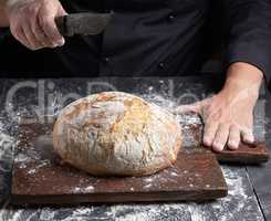 male chef hand holds knife over a whole round baked loaf of brea