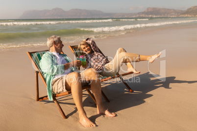 Senior couple having cocktail drink while relaxing on sun lounger at beach