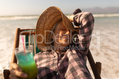 Senior woman having cocktail drink while relaxing on sun lounger at beach