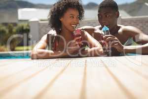 Happy young African American couple leaning on edge of pool and drinking cocktail