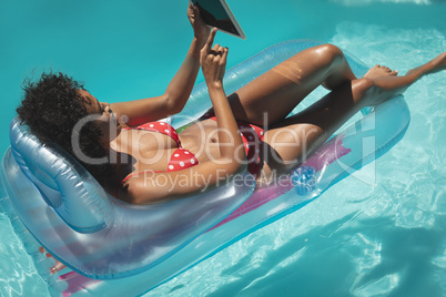 Young African American woman relaxing on pool lounger and using digital tablet