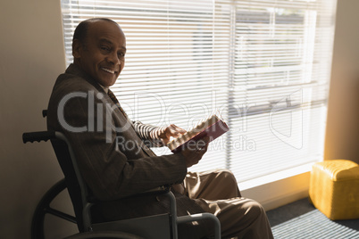Side view of disable senior man with book sitting on wheelchair and looking at camera