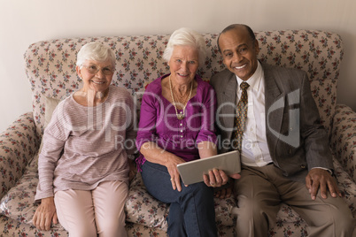 Front view of senior friends with digital tablet sitting on sofa and looking at camera