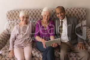 Front view of senior friends with digital tablet sitting on sofa and looking at camera