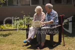 Front view of senior couple sitting on bench and using laptop in garden