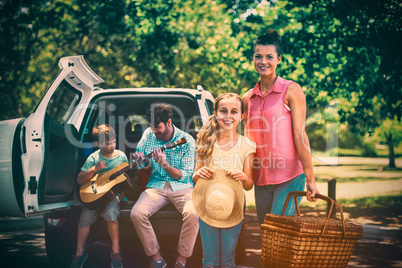 Portrait of mother and daughter standing with picnic basket while father and son playing guitar in b