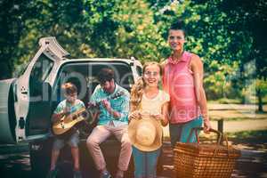 Portrait of mother and daughter standing with picnic basket while father and son playing guitar in b