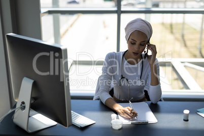 Beautiful mixed-race female doctor talking on mobile phone while writing on clipboard