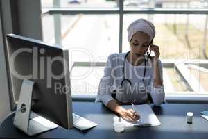 Beautiful mixed-race female doctor talking on mobile phone while writing on clipboard