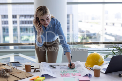 Blonde Caucasian female architect talking on mobile phone while working at desk in office