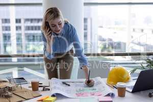 Blonde Caucasian female architect talking on mobile phone while working at desk in office
